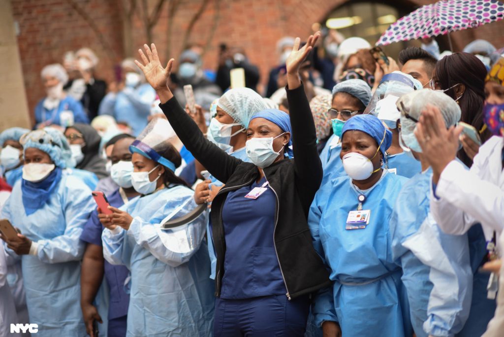 A group of cheering nurses. The nurses are wearing protective gear like masks, scrubs, and medical gowns.