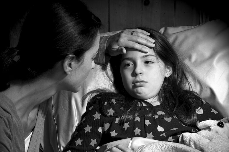 A little girl is in bed. A woman feels her forehead to see if she has a fever.